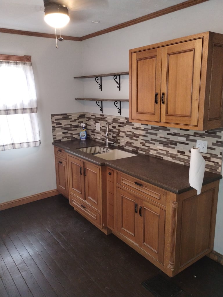 Modern kitchen with wooden cabinets, dark countertop, gray tile backsplash, stainless steel sink, floating shelves, and pendant lighting