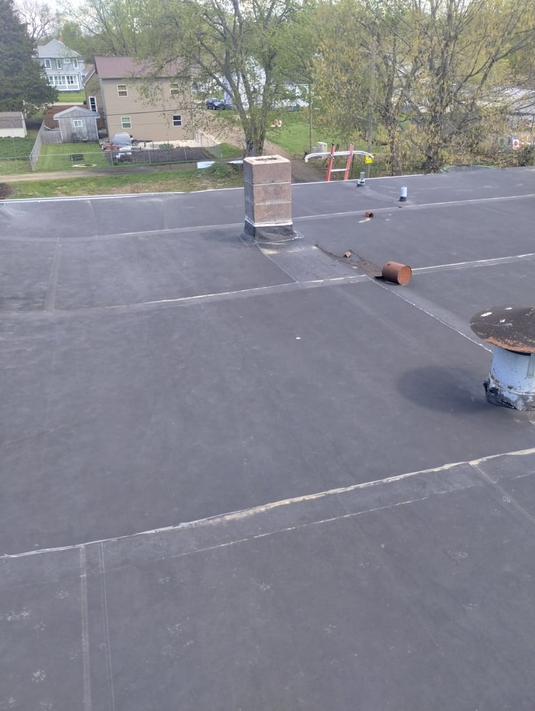 Flat roof with brick chimney, metal piping, and residential house with trees in background