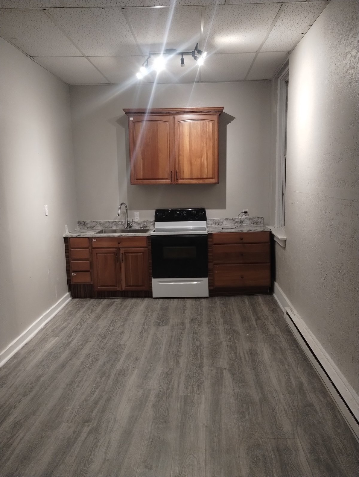 Unfinished kitchen space with wooden cabinetry, black and white stove, gray vinyl flooring, and white drywall walls with track lighting overhead