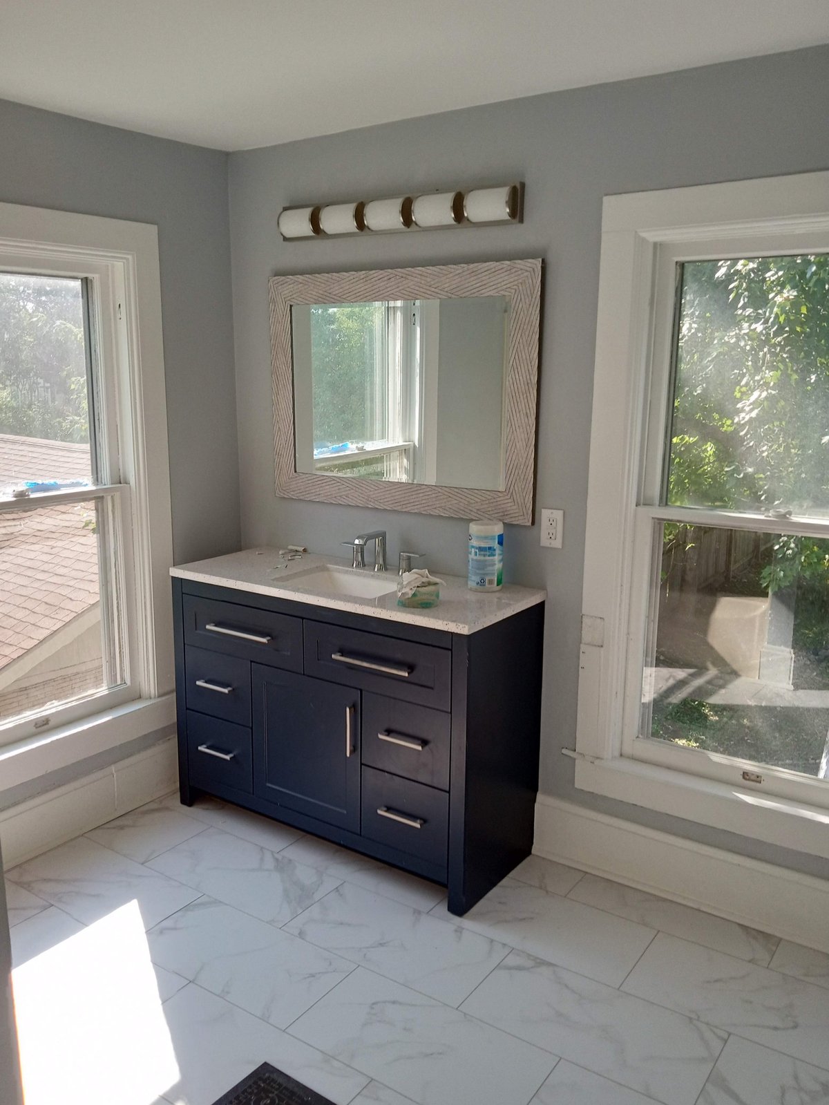Modern bathroom with navy vanity, white quartz countertop, framed mirror, decorative wall sconces, and large windows overlooking green landscape