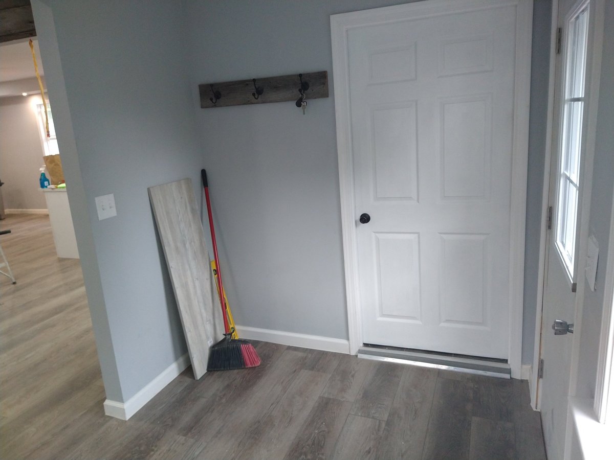 Mudroom corner with white closet door, dark wood coat rack, cleaning supplies, light gray walls, and laminate flooring