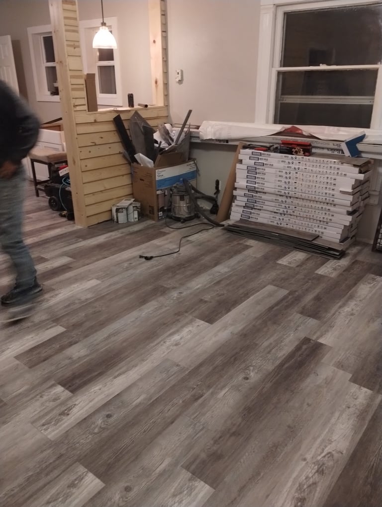 Cluttered room with gray wood-look flooring, cardboard boxes, stacked items, and moving supplies during home renovation or relocation