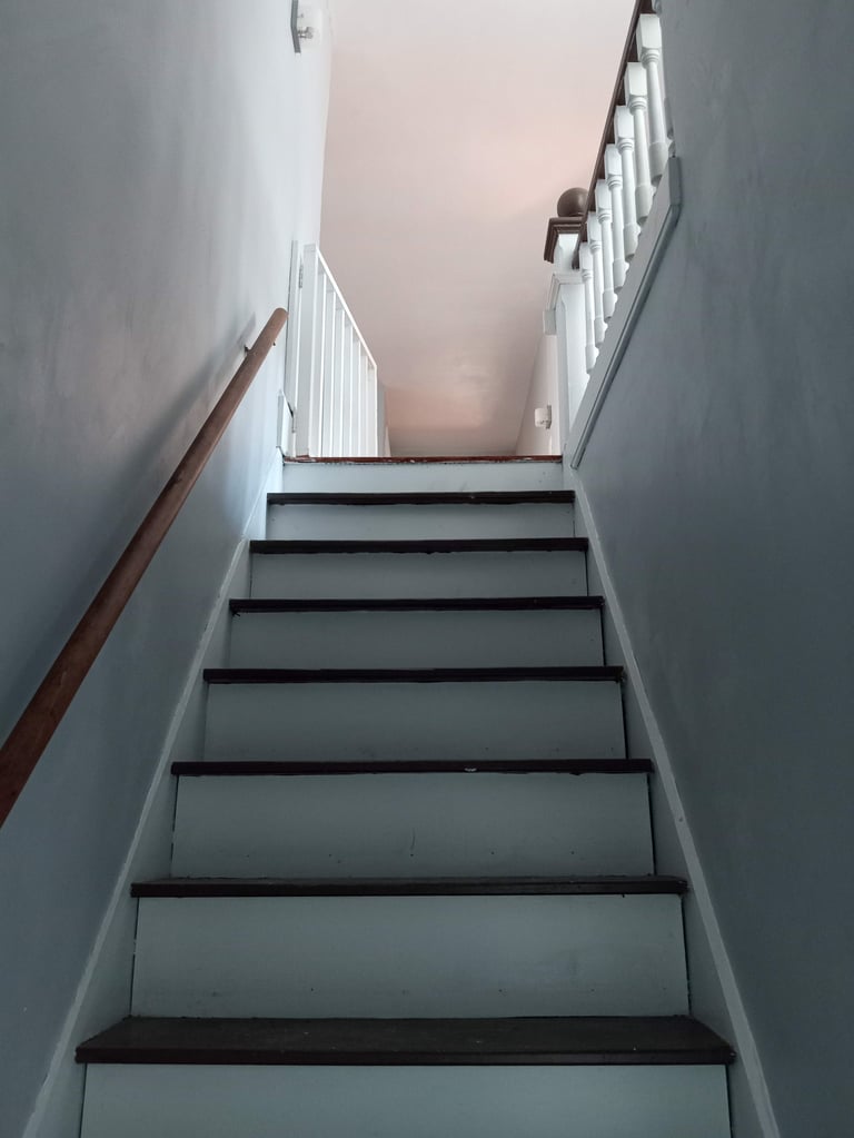Staircase with gray steps and brown handrail ascending toward a white landing with railings and bright ceiling light above