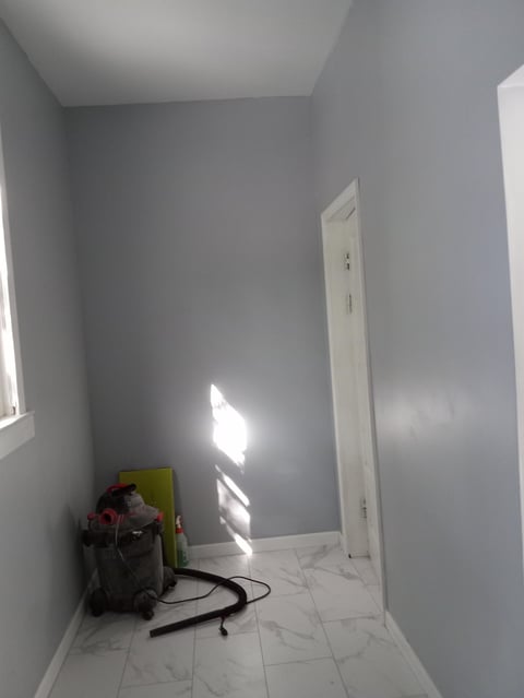 Empty room with gray walls, white tile floor, sunlight from window, and construction materials in corner