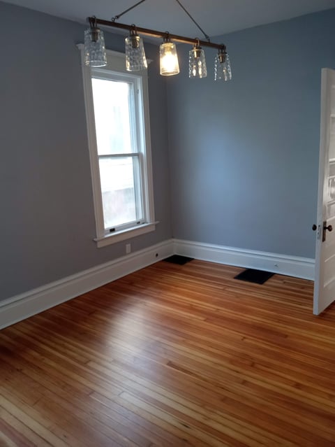 Empty bedroom with blue walls, wooden floor, window, glass pendant light fixture, and white trim