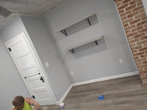 Empty room with gray walls, two floating shelves, white door, exposed brick wall, and wood laminate flooring