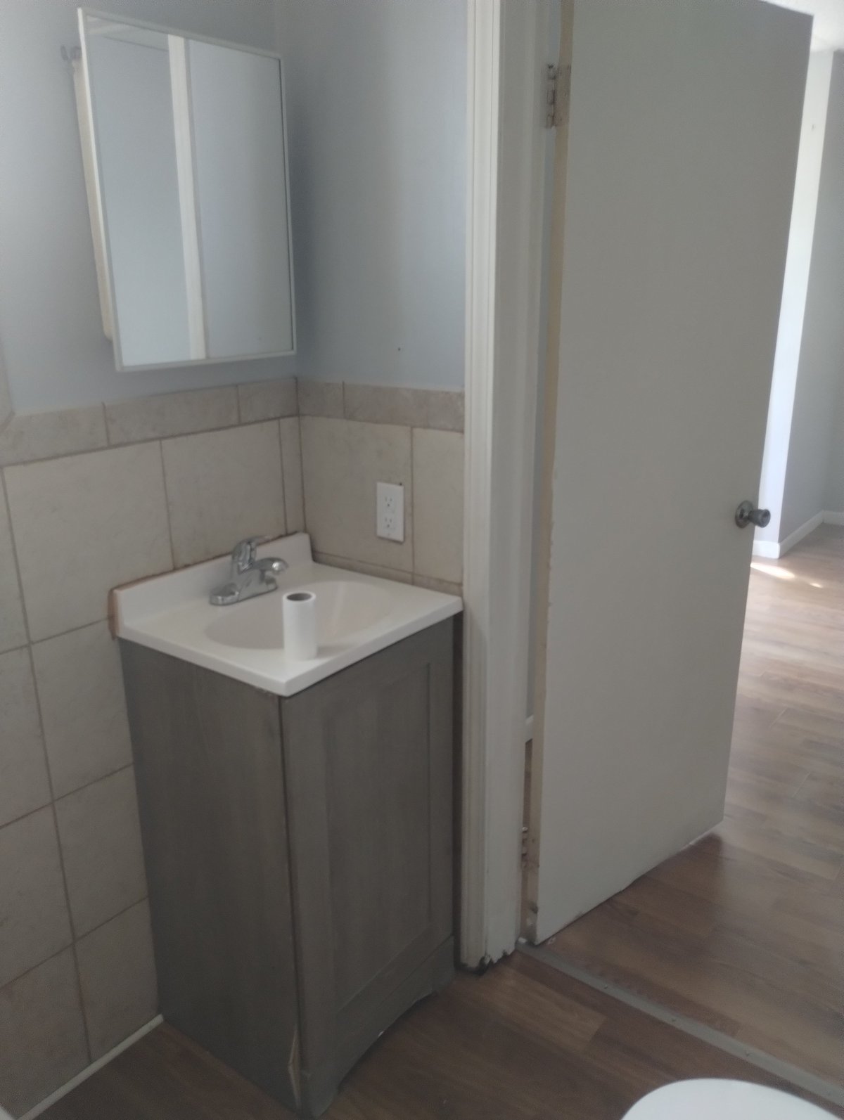 Compact bathroom corner with small gray vanity, white sink, mounted mirror, and beige tile wall