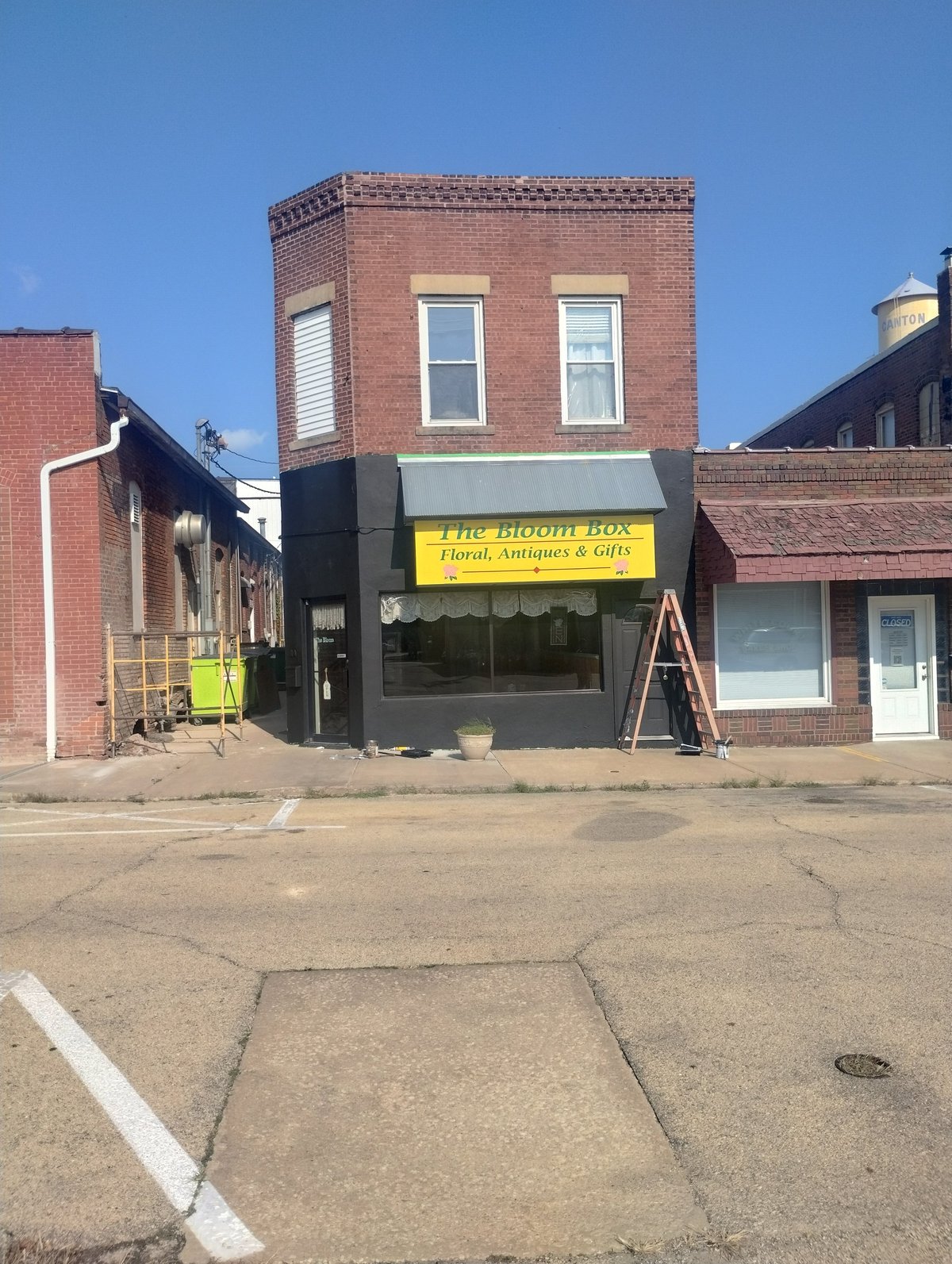 Street view of The Bloom Box florist storefront in a red brick building with yellow signage under clear blue sky