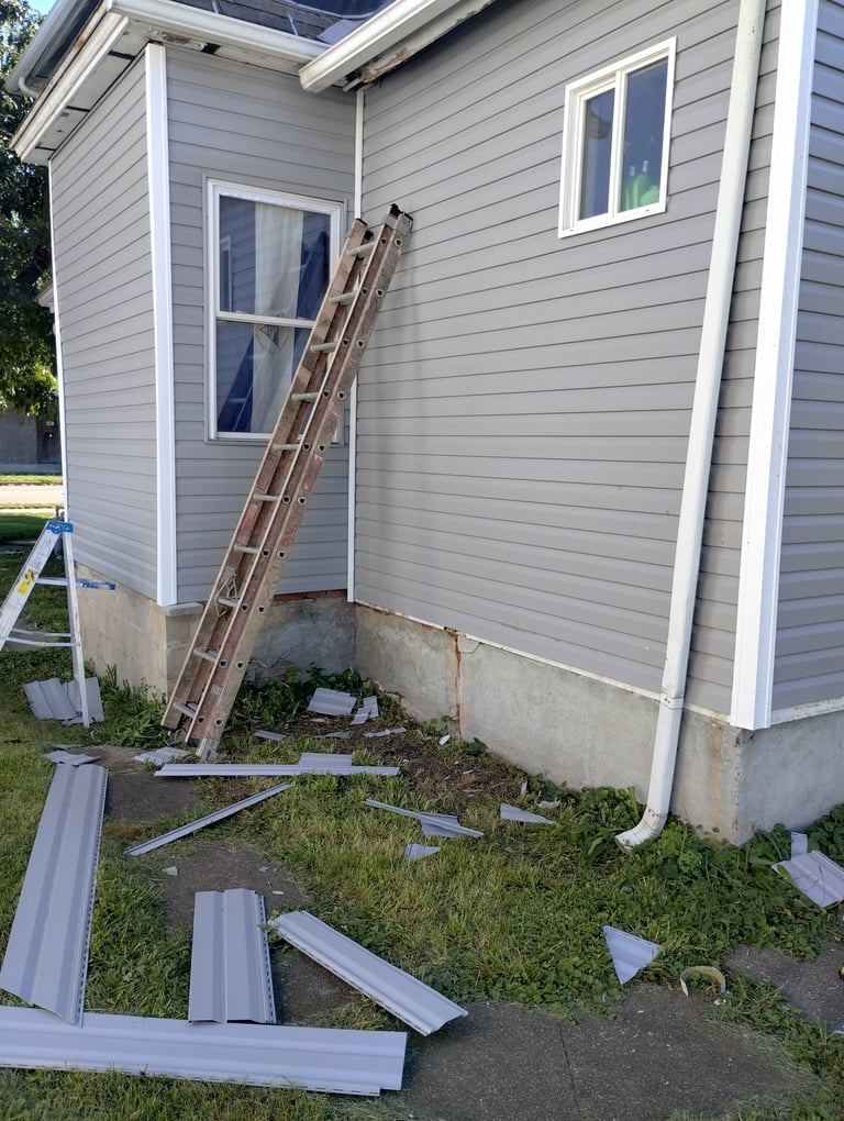 Wooden ladder leaning against gray house siding with construction materials and concrete foundation visible in yard