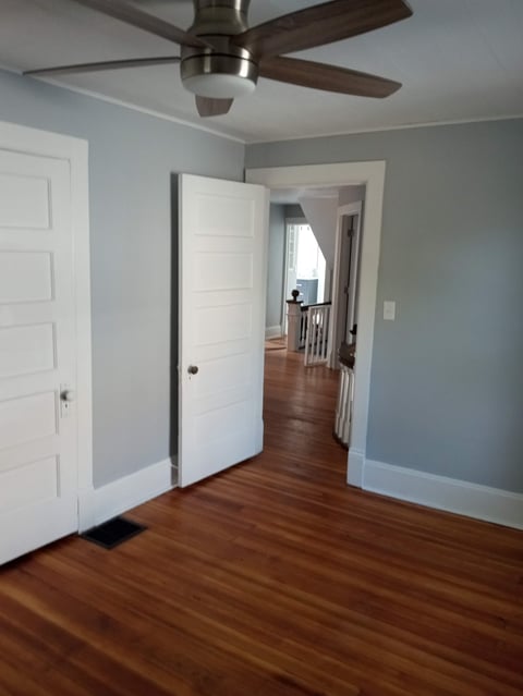 Spacious bedroom with hardwood floors, white doors, light blue walls, ceiling fan with recessed lighting, and views into hallway