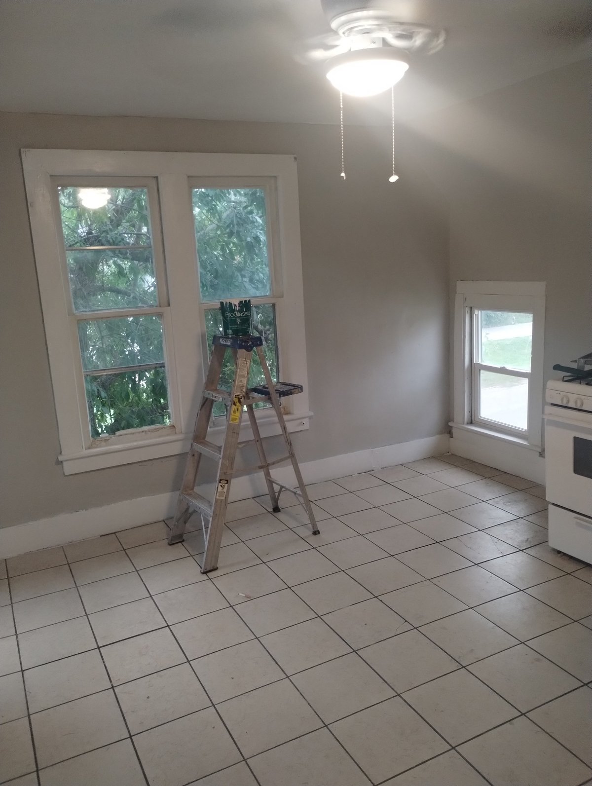 Empty kitchen with beige walls, white tile floor, ladder against large window, ceiling light fixture, and white appliances