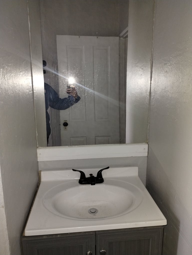 Bathroom mirror reflection showing a person taking a selfie, white sink with black faucet, and gray vanity cabinet