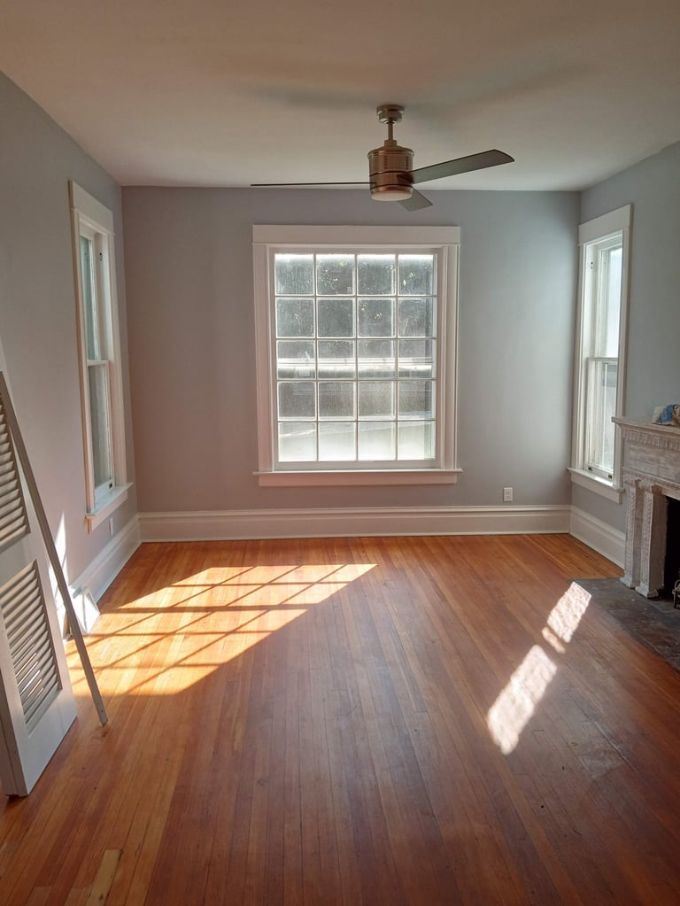Empty bedroom with hardwood floors, large central window, bronze ceiling fan, and pale pink accent wall