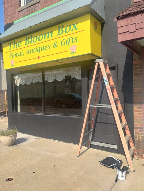 The Bloom Box storefront with yellow signage, featuring a wooden ladder and paint supplies on the sidewalk during renovation