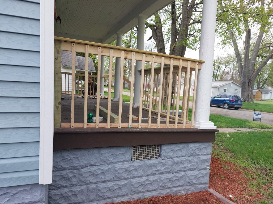 Front porch with new wooden railing framework under construction on a light blue house with blue stone skirt, white columns, and trees in the background