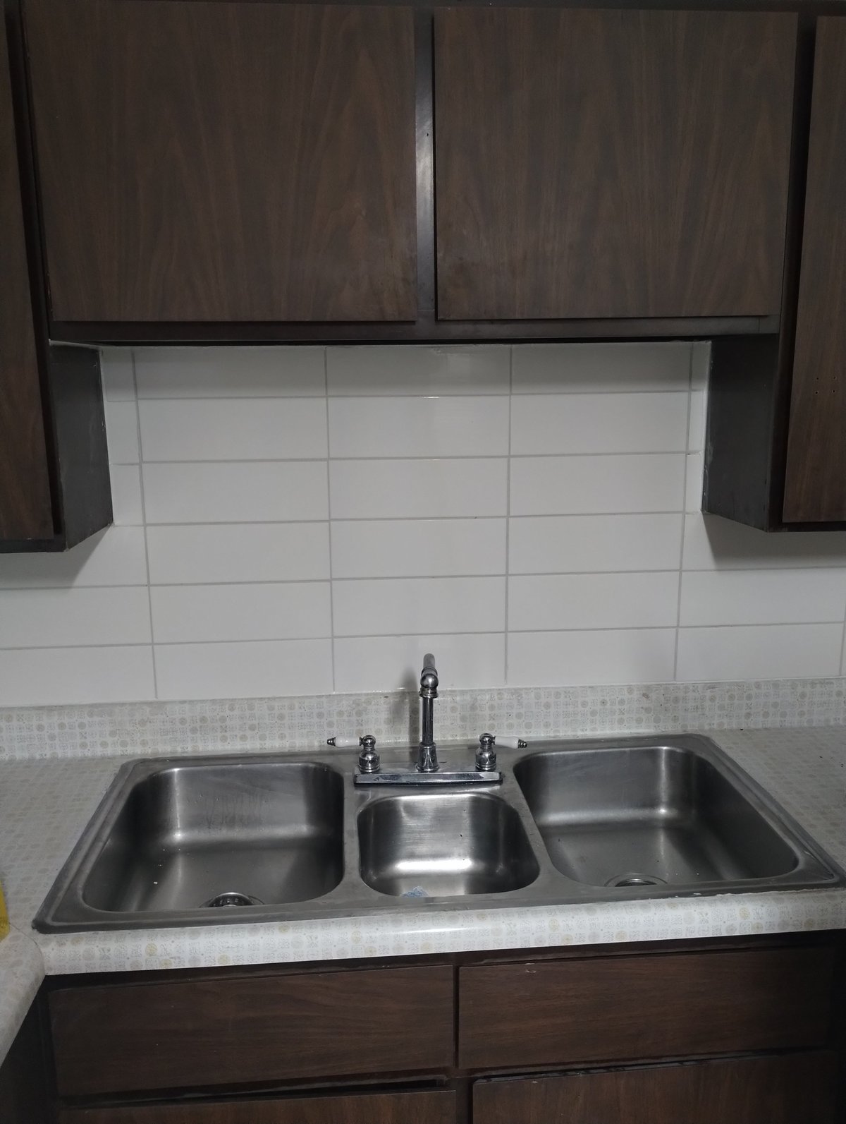 Modern kitchen sink with three stainless steel compartments and faucet, white tile backsplash, dark wood cabinets above