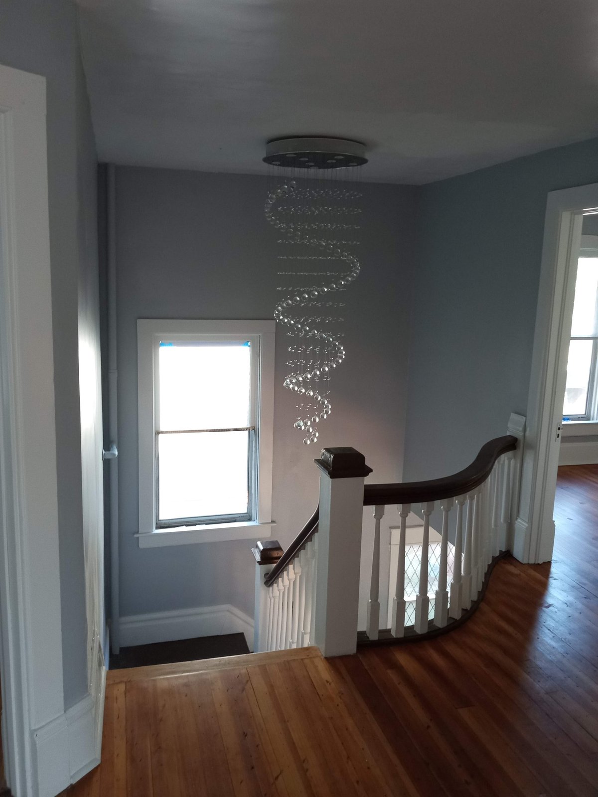 Stairwell landing with spiral crystal chandelier, wooden banister, hardwood floors, and door leading to room with windows