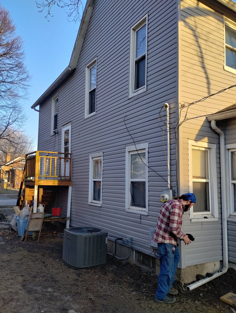 Gray two-story house with yellow deck addition. A person in a red plaid shirt and blue cap stands outside. Bare trees and clear sky visible.