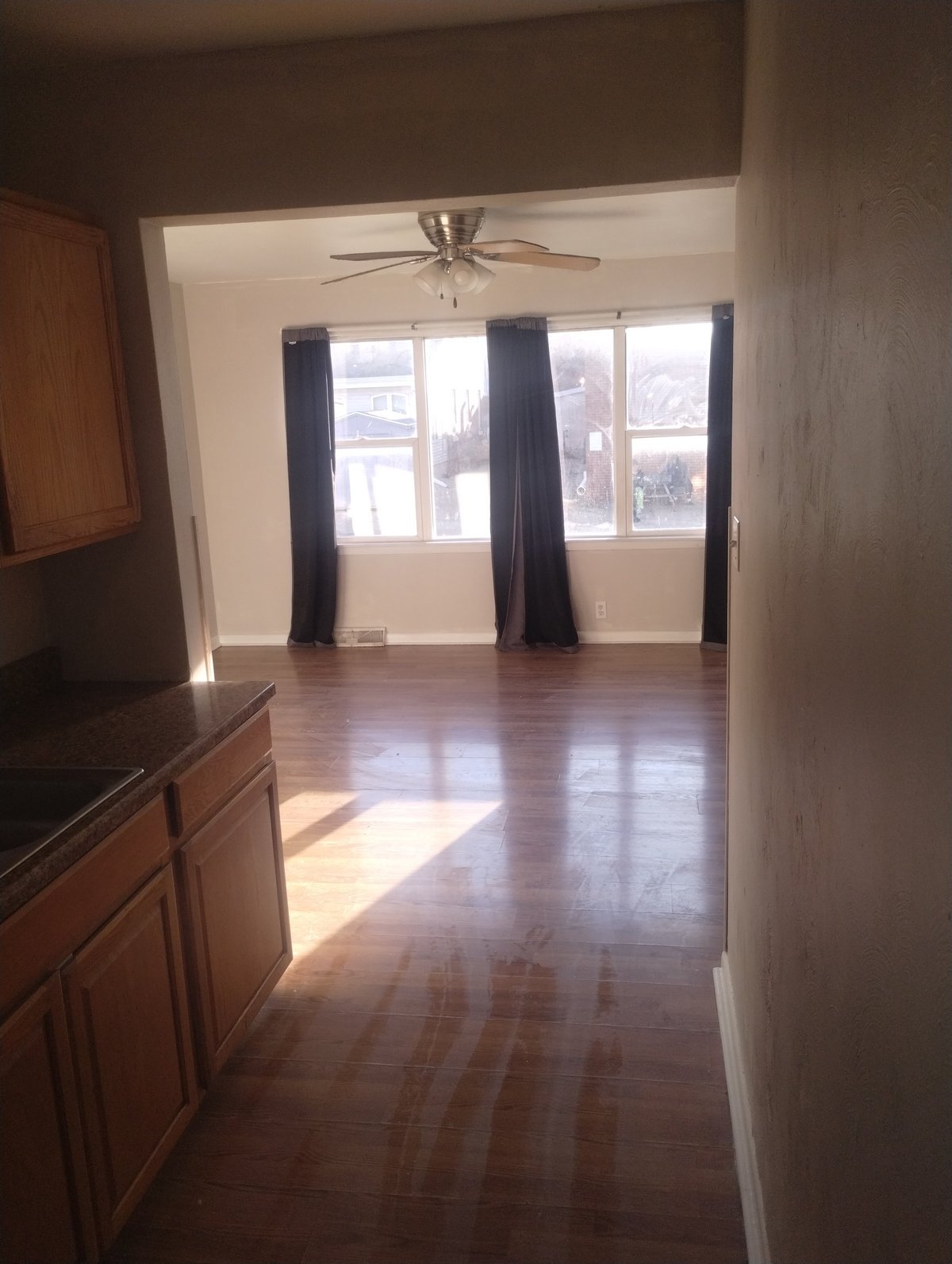 Spacious apartment interior with hardwood flooring, dark curtains on large windows, ceiling fan, and wood cabinetry visible in foreground