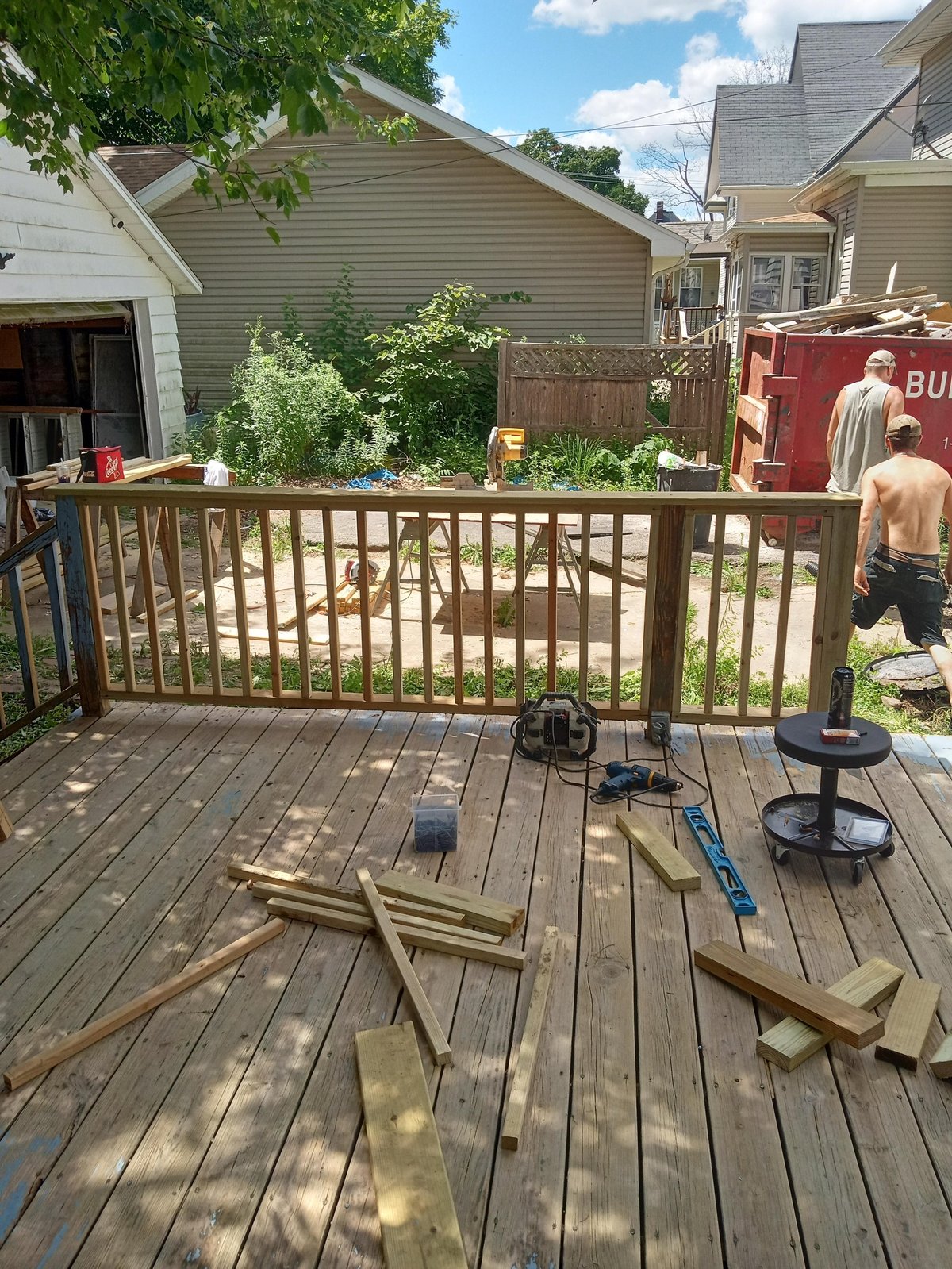 Wooden deck with brown railing under construction, tools and materials scattered on boards, residential houses and yard visible in background