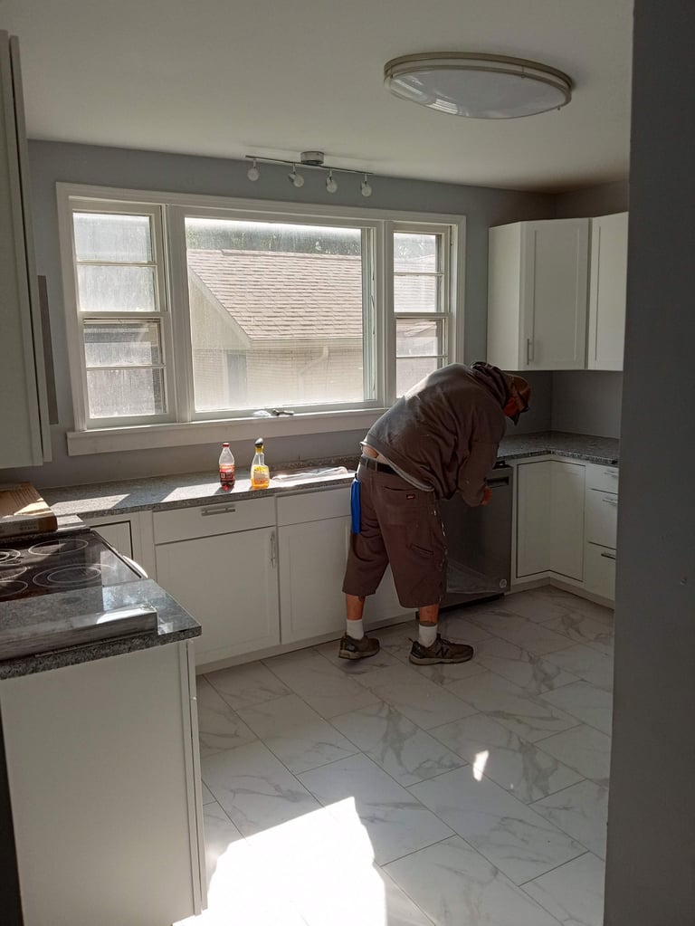 Worker in brown jacket bending over while working in a modern kitchen with white cabinets and marble flooring