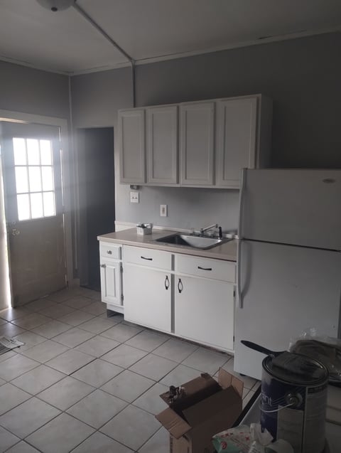 Small kitchen with white cabinets, sink, stovetop, and refrigerator. Tile floor and exit door visible on the left.