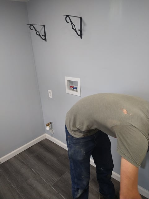 Person bending down to inspect wall outlet in gray room with shelving brackets and small picture frame visible