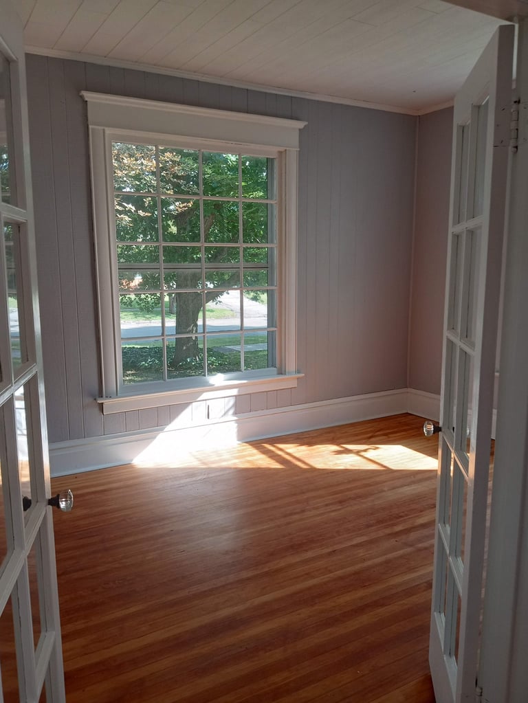 Empty room with hardwood floors, mauve walls, and large window overlooking trees and driveway