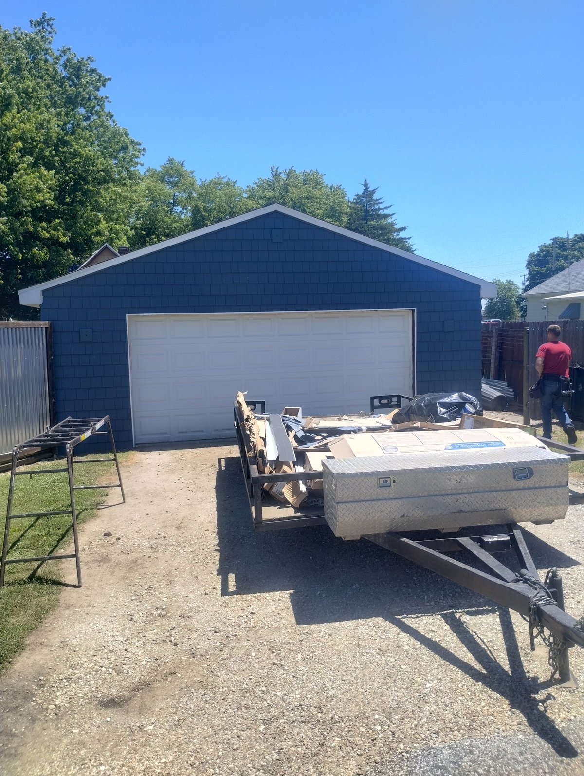 Blue garage with white garage door and a utility trailer parked in front on gravel driveway