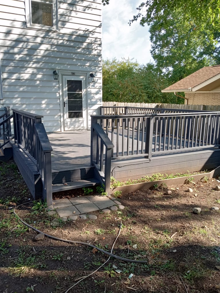 Residential deck with dark railing and composite boards, white ramp with slope leading to side of house, surrounded by green trees