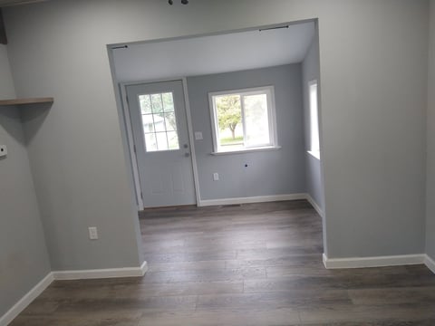 Empty room with hardwood flooring, white walls, and entryway with glass door and windows overlooking outdoor landscape