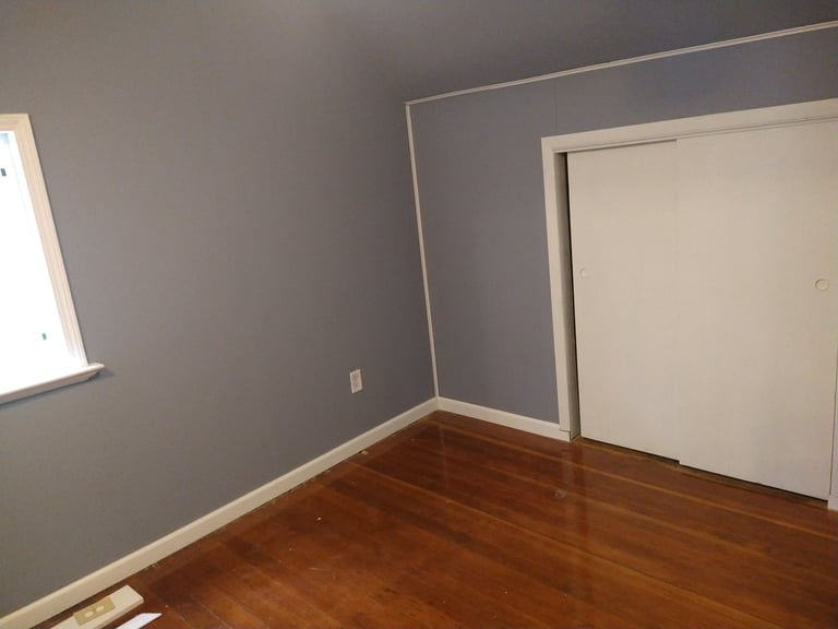 Empty room with gray walls, hardwood floor, window on left, and closed white door on right