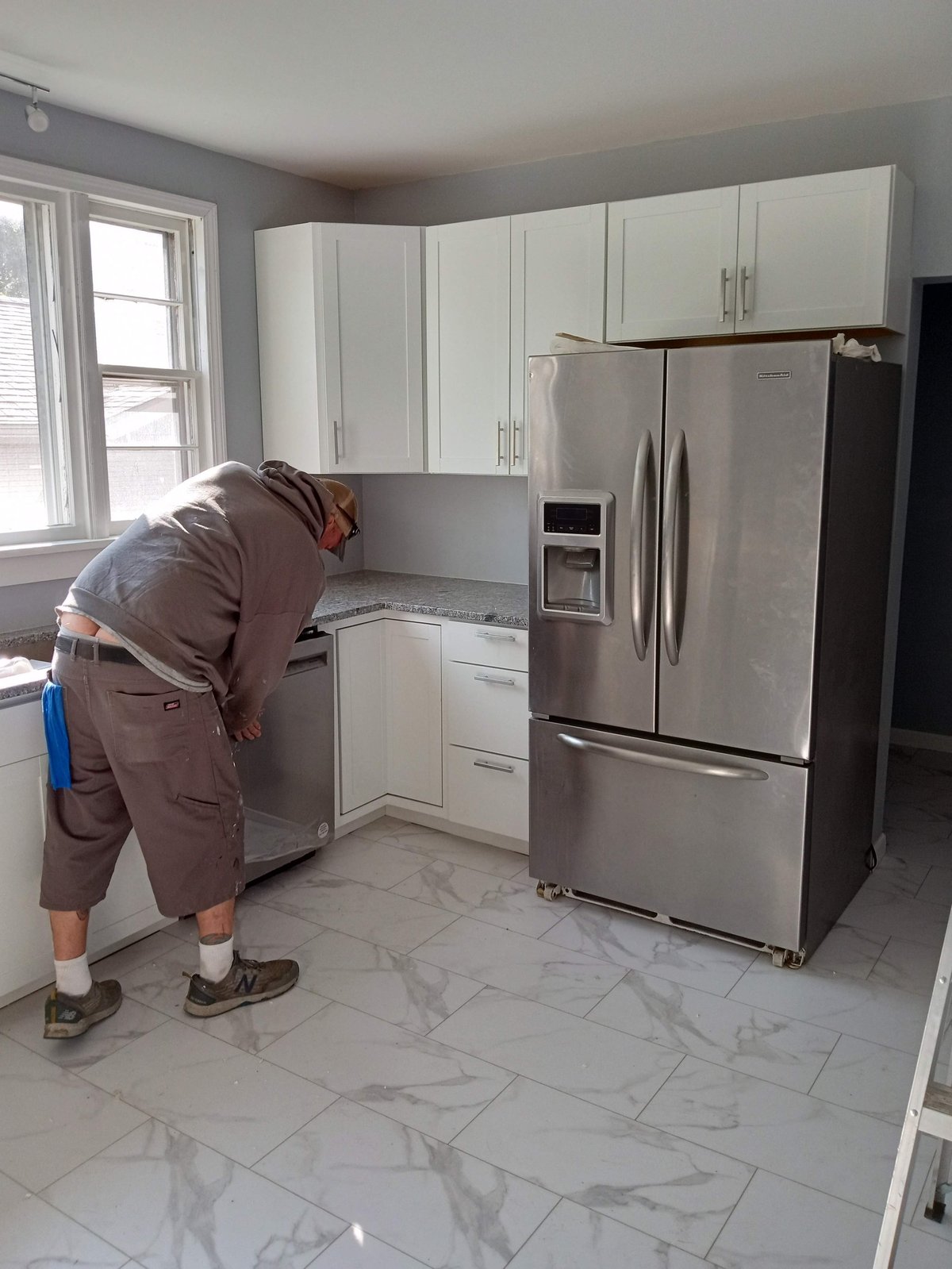 Worker in brown uniform leaning over kitchen counter in modern kitchen with white cabinets and stainless steel refrigerator