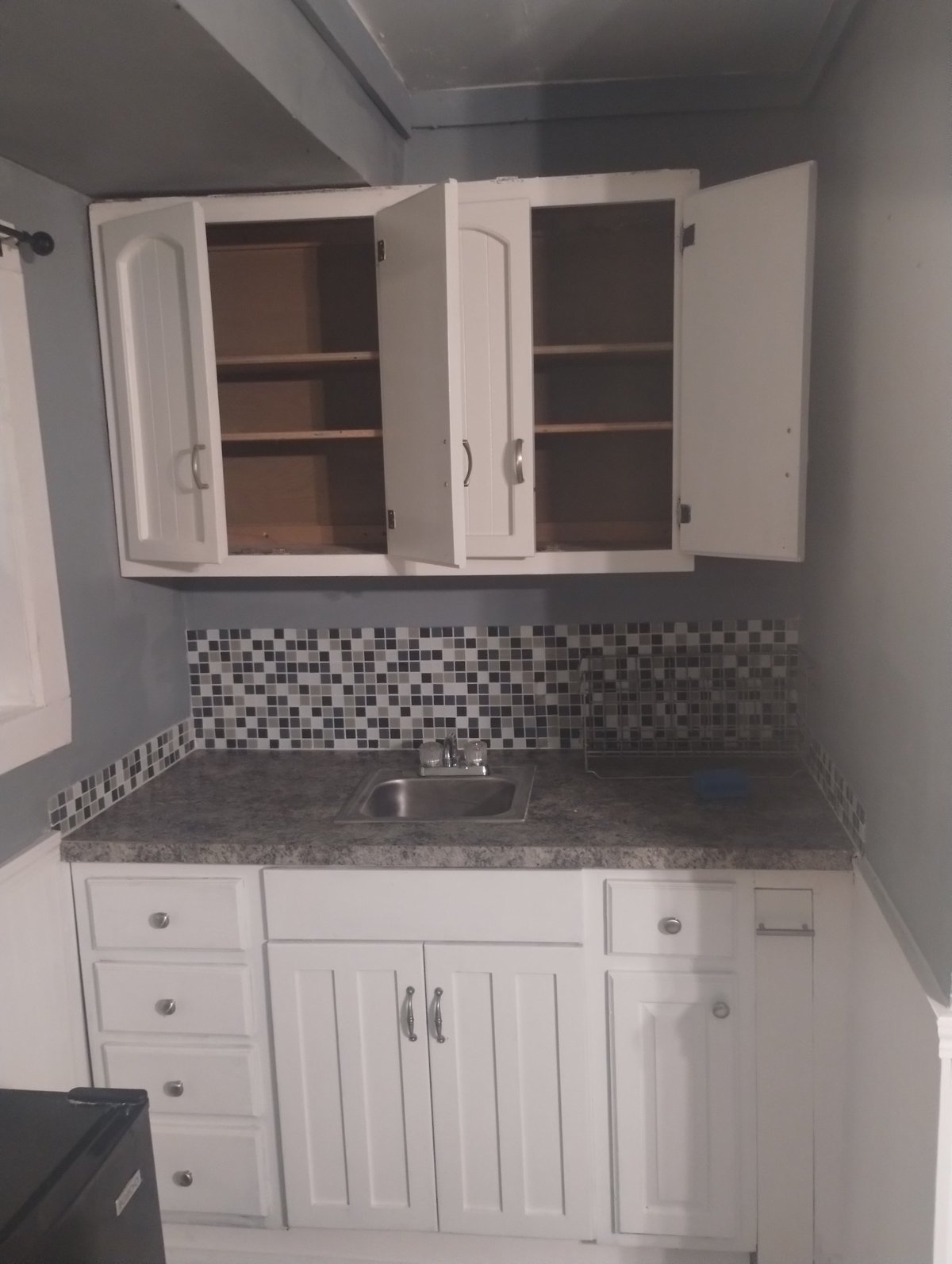 Small kitchenette with white cabinetry, dark gray countertop, stainless steel sink, and black and white checkered tile backsplash