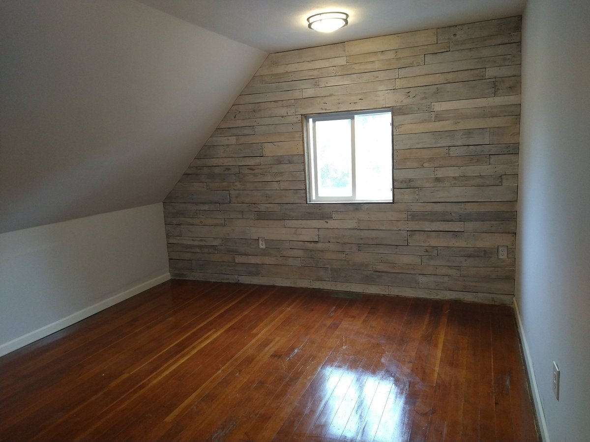 Empty attic room with wood plank accent wall, hardwood floor, rectangular window, and ceiling light fixture