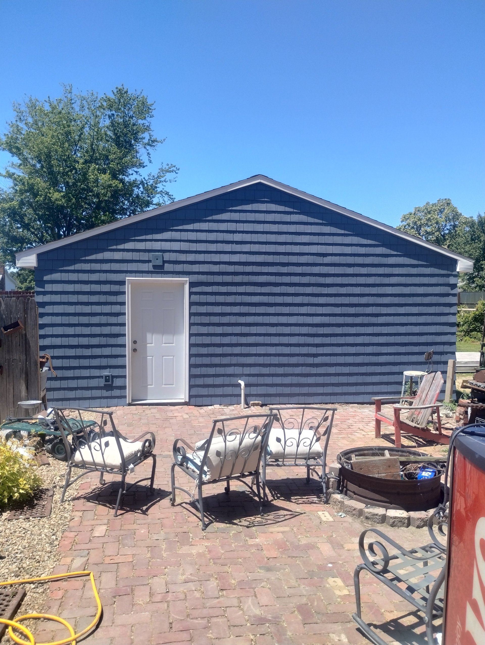 Blue shed with white door on brick patio, metal chairs and trees under clear sky