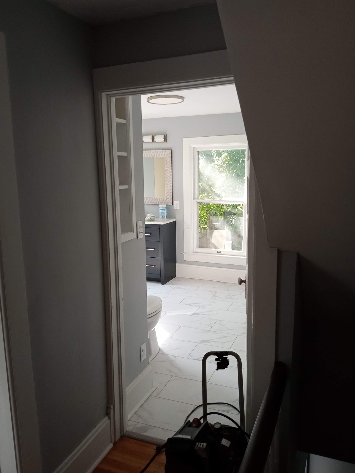 View through doorway into a bright modern bathroom with white tile flooring, dark vanity, and window overlooking greenery
