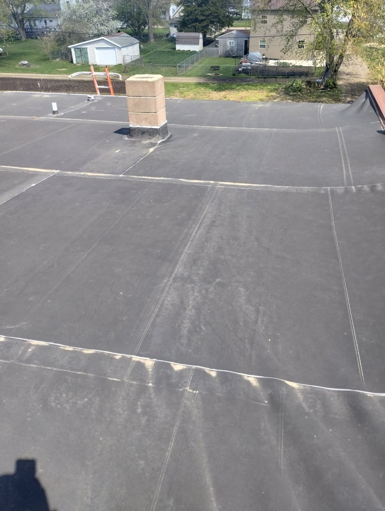 Flat roof under construction with seams and material laid out, houses and storage sheds visible in background