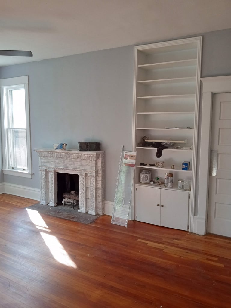 Empty living room with hardwood floor, brick fireplace, white built-in shelving unit, window, and beige walls