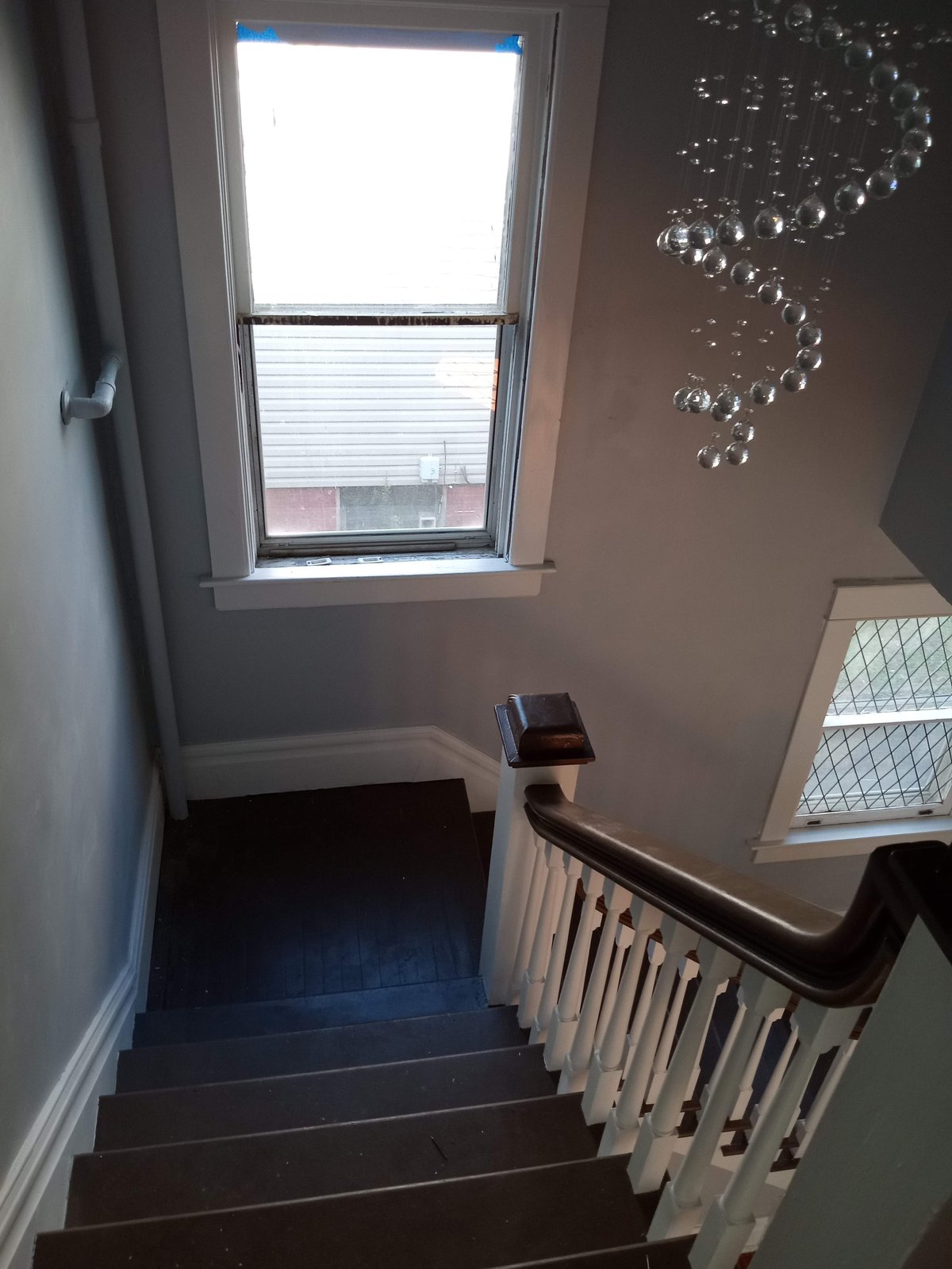 Dark wooden staircase with white railings descending toward a window with blinds, white walls, and a crystal chandelier overhead