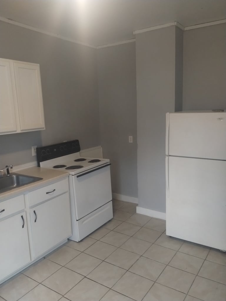 Small kitchen with white appliances, gray walls, white cabinetry, tile flooring, and a black chair near the stove