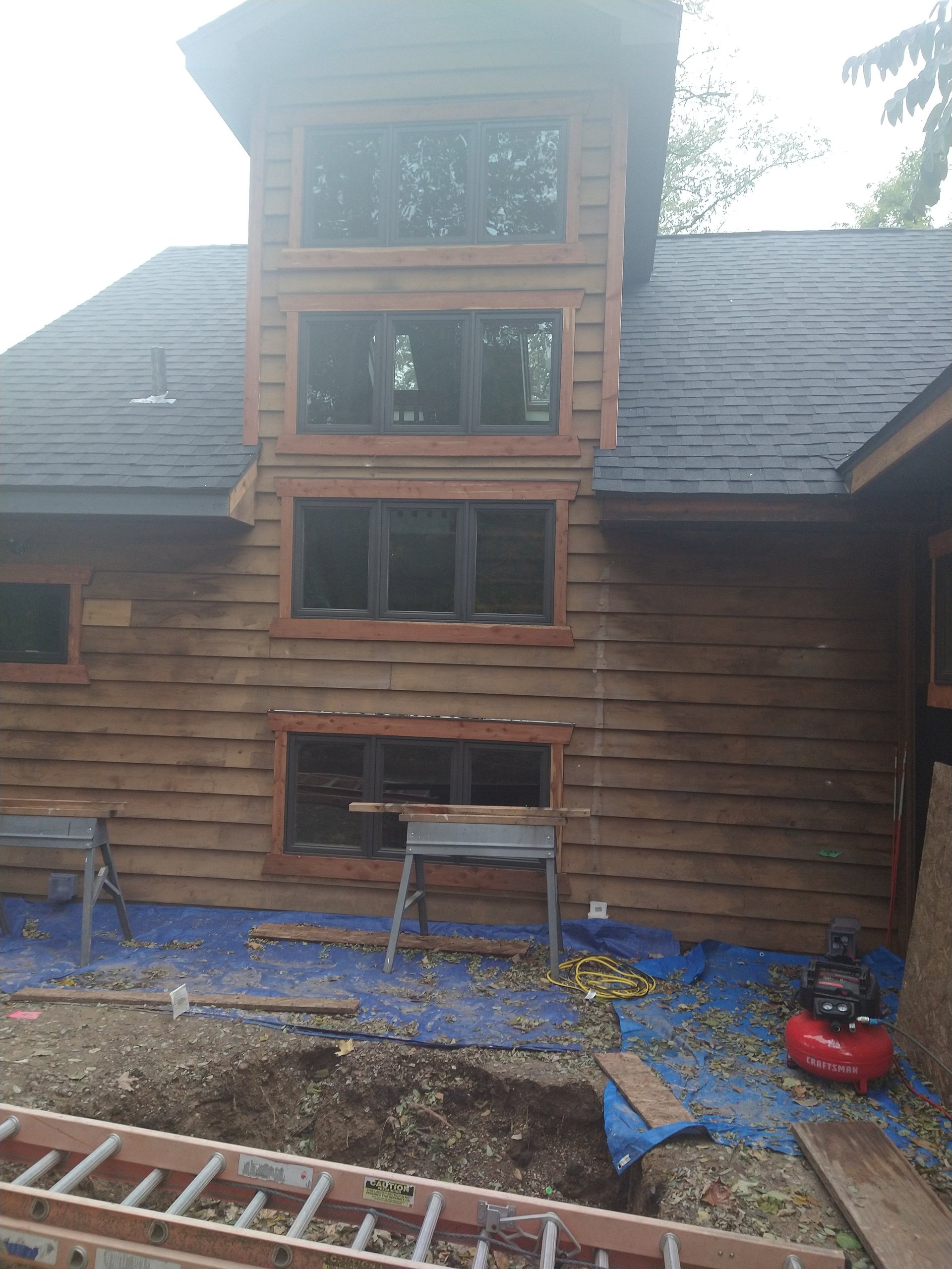 Home under construction with wooden cedar shake siding, stacked replacement windows, blue tarps, and construction equipment in yard