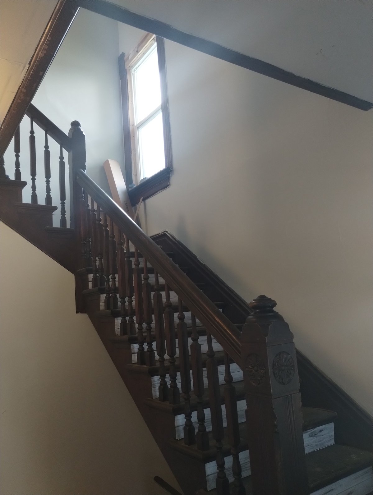 Dark wooden staircase with railings ascending toward a window with bright light streaming through an upper level