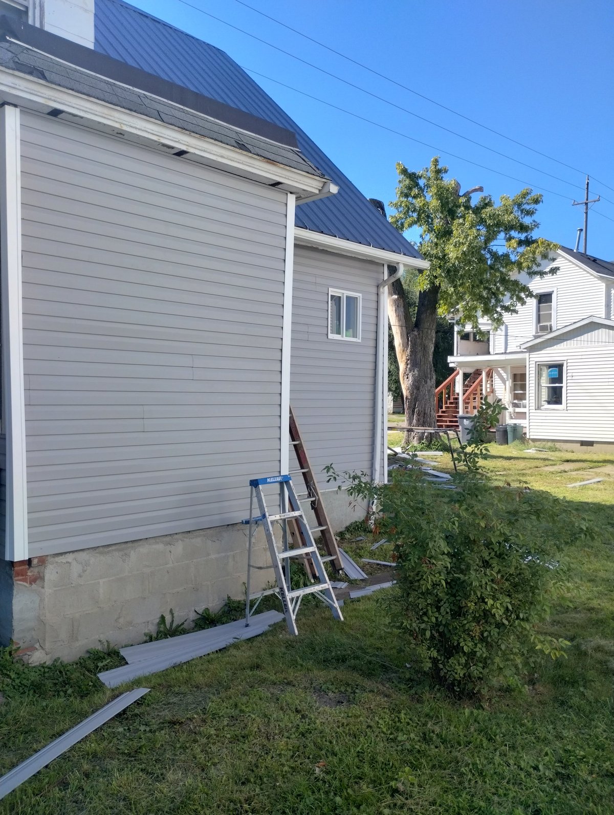 White house addition with blue metal roof, ladder against side, and neighboring homes visible in yard setting