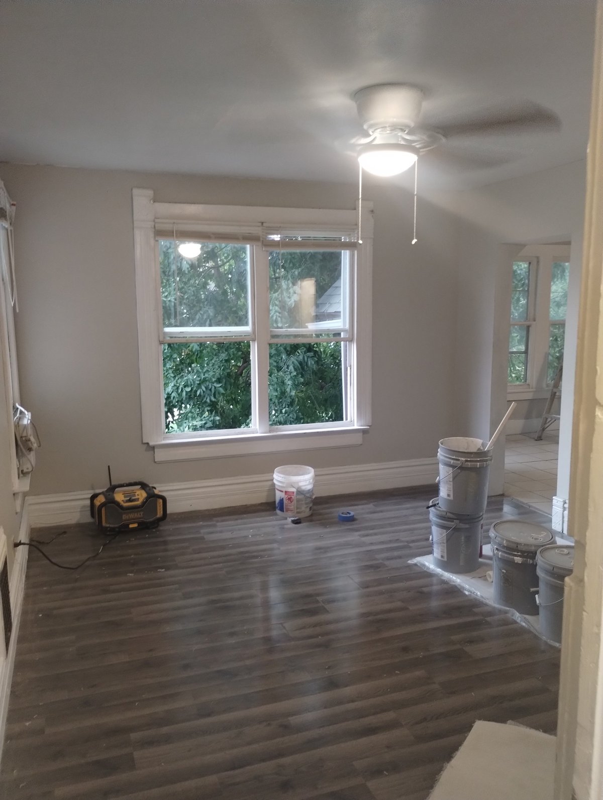 Empty room under renovation with dark hardwood flooring, white ceiling fan, large window overlooking trees, and construction materials