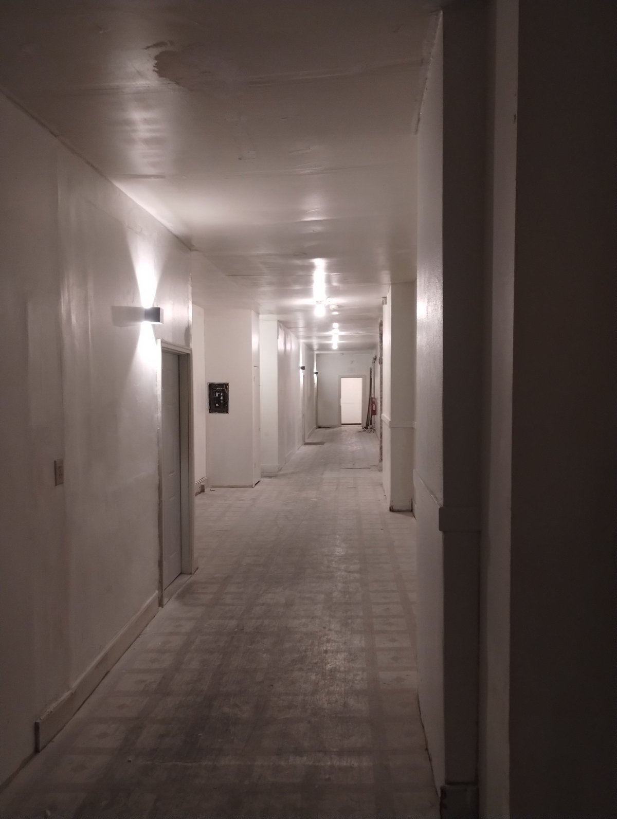 Long empty hallway with white walls, fluorescent ceiling lights, tiled floor, and columns, perspective extending into distance