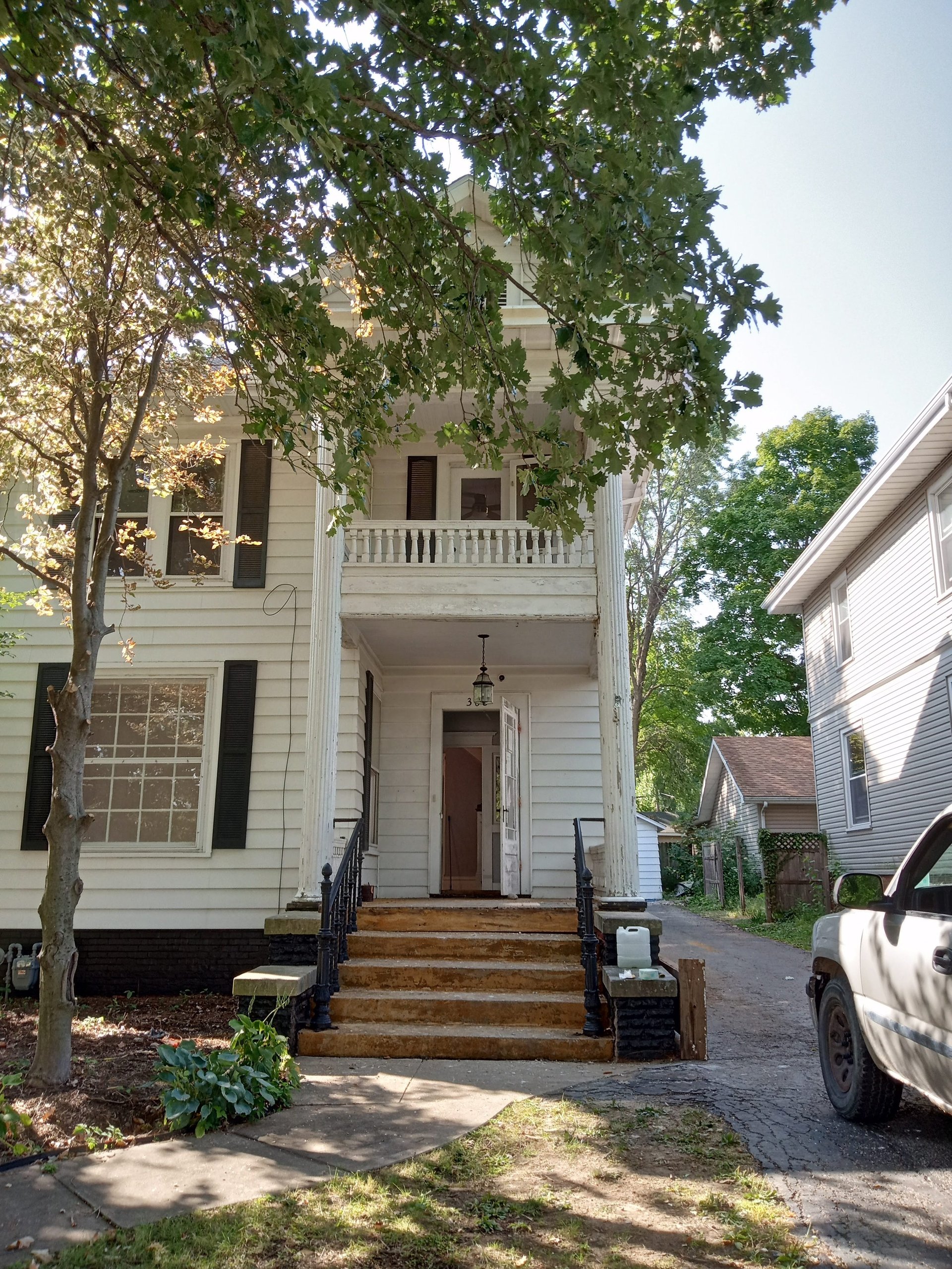 White two-story colonial-style house with red front door, wooden porch steps, and ivy-covered tree in front driveway