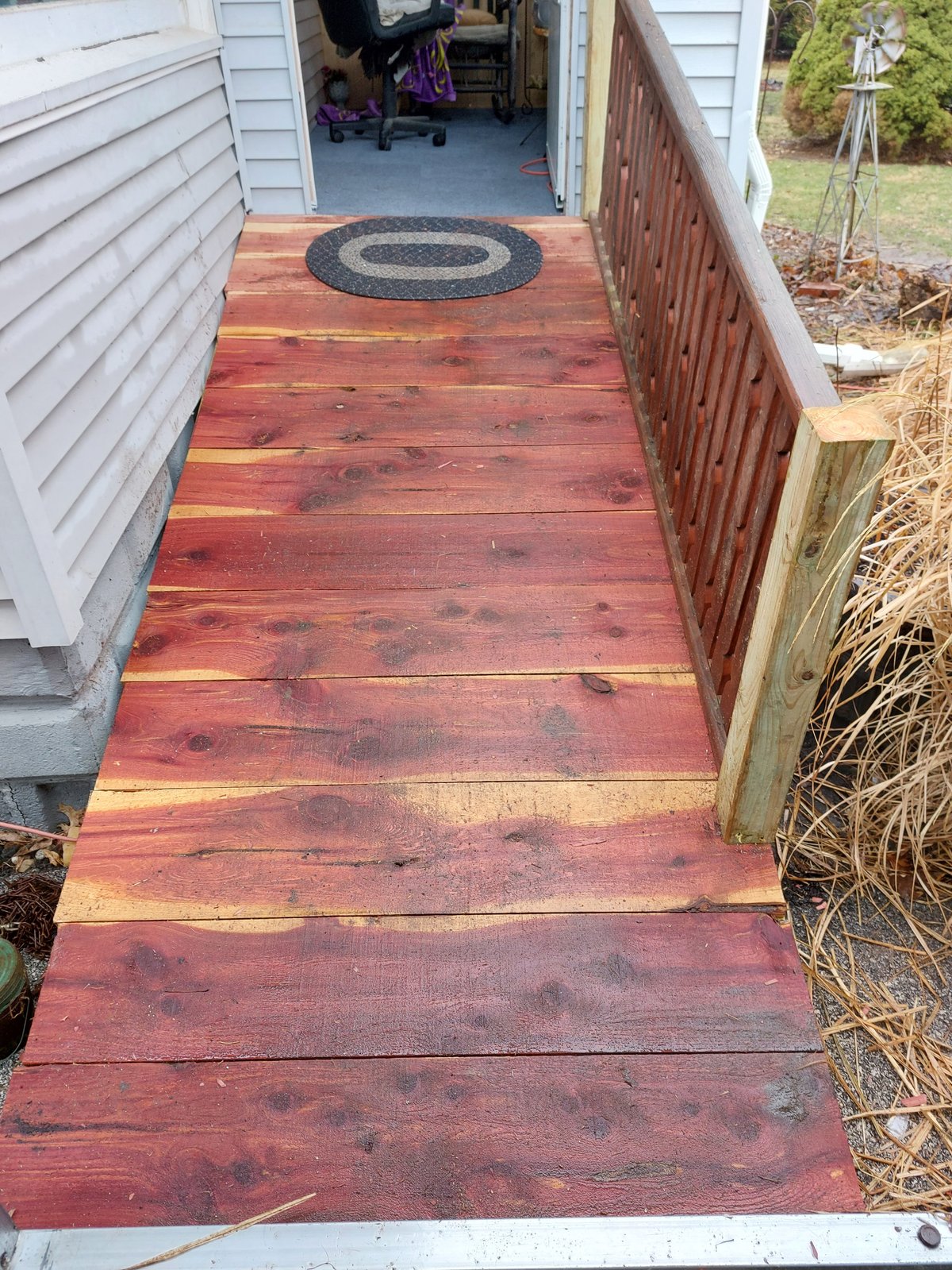 Wooden deck with rope entrance mat leading to garage doorway, flanked by white siding and brown railing