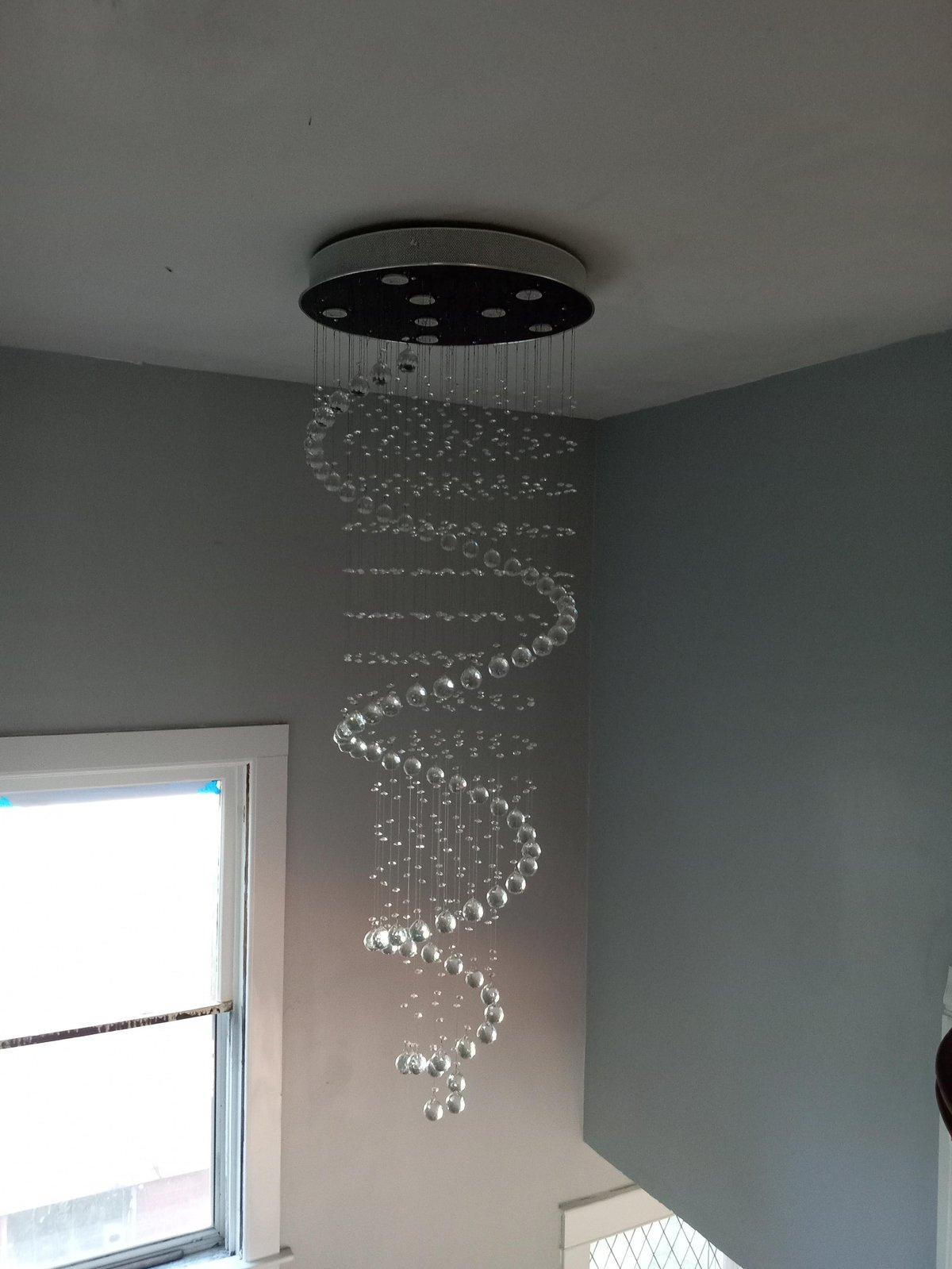 Modern spiral crystal chandelier hanging from black ceiling fixture in grey room with window