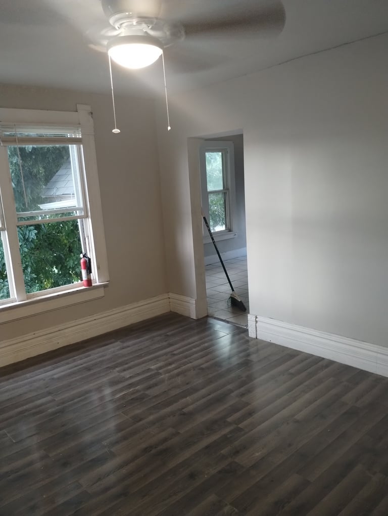 Empty room with hardwood flooring, white walls, ceiling fan with light fixture, window on left, and doorway to adjacent room